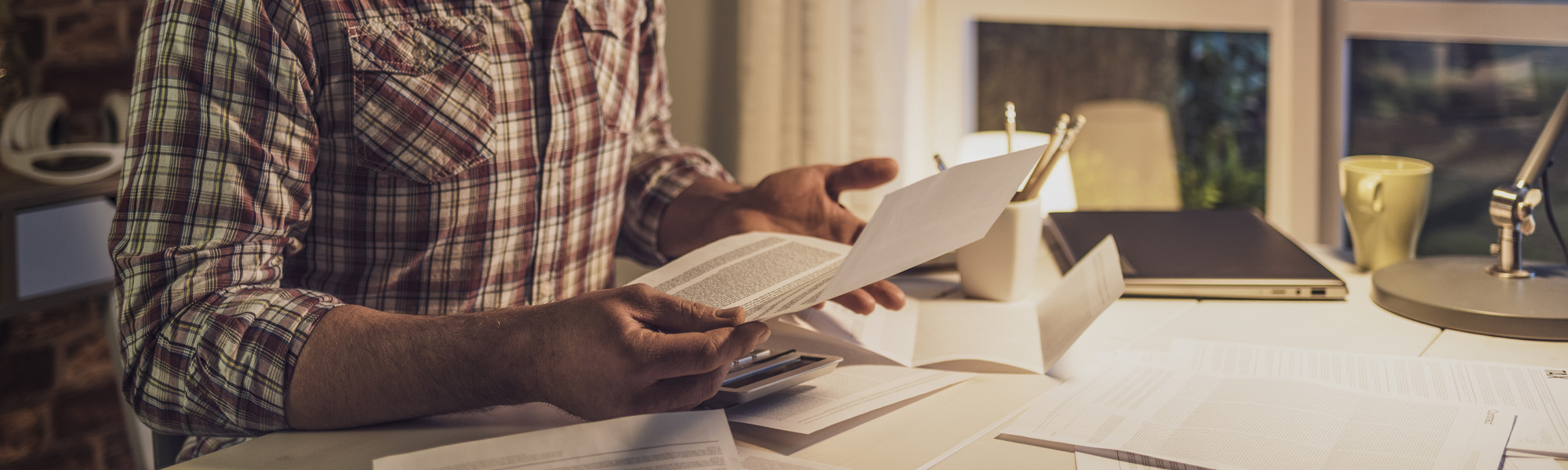 A person in a plaid shirt reviews documents related to California debt collection harassment cases at a desk strewn with papers, near a laptop and a lamp, suggesting a scene of late-night work or study