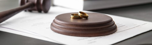 A pair of wedding rings rests atop legal documents related to domestic identity theft next to a judge's gavel, suggesting the context of a divorce or marriage dissolution proceeding.