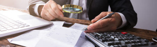 A meticulous financial analysis of fraudulent credit reporting: business person scrutinizing receipts with a magnifying glass while taking notes and using a calculator next to a laptop.