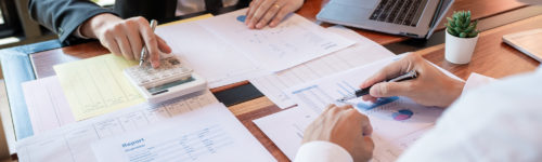 Two professionals analyzing financial charts and using a calculator on a wooden desk with a laptop in the background, concerned about inaccurate account information.