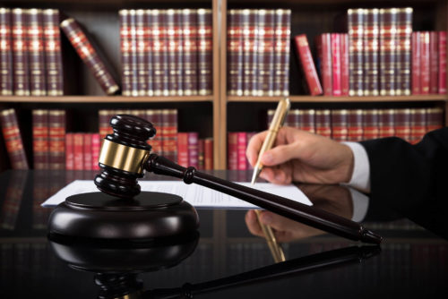 A judge's gavel rests on a sounding block with a person writing on a legal document in the background, symbolizing the administration of justice within a law library setting.