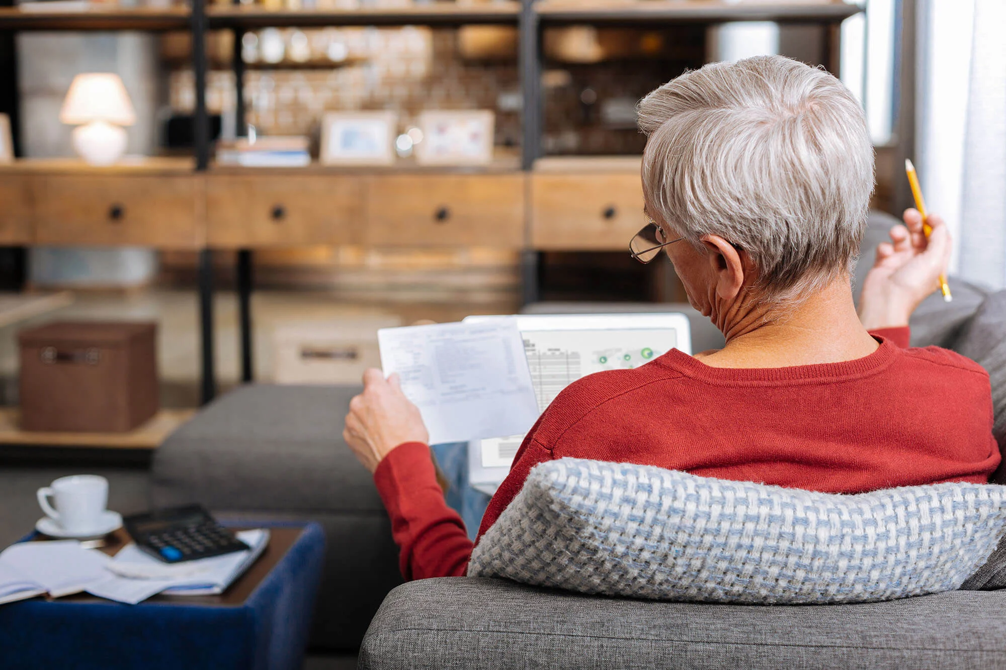 A person with short hair wearing a red sweater and glasses is sitting on a blue couch, holding a document and a pencil, apparently reviewing or working on paperwork, with a laptop and a cup of coffee on the table in the background, suggesting a focused and cozy home office setting.