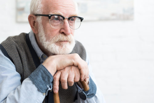A senior gentleman with a beard, wearing glasses and a vest, resting his chin on his hands which are gripping a walking stick, looking thoughtfully into the distance.