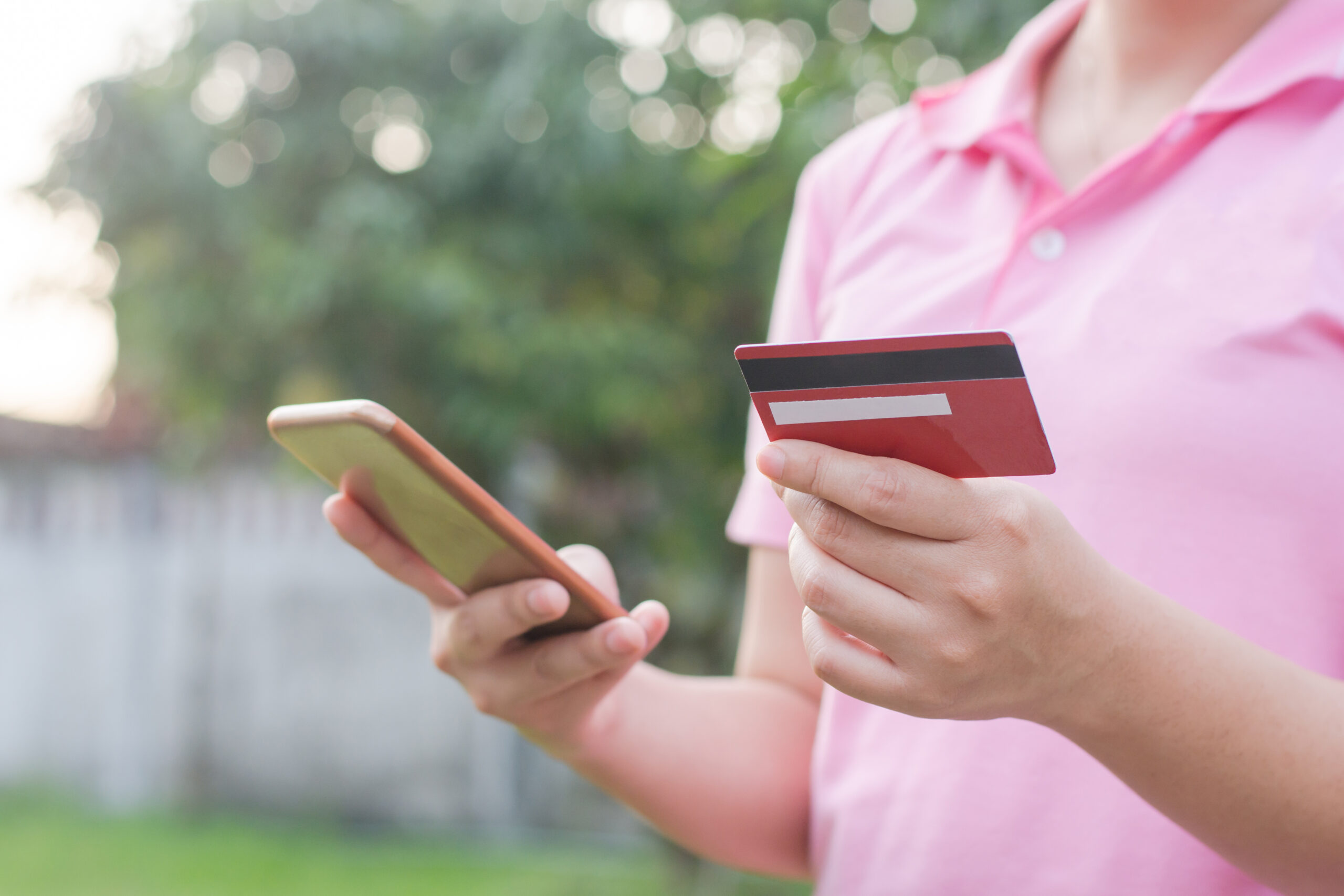 Mobile payment: a person in a pink shirt holding a smartphone in one hand and a credit card in the other, possibly making an unauthorized online transaction or managing finances.