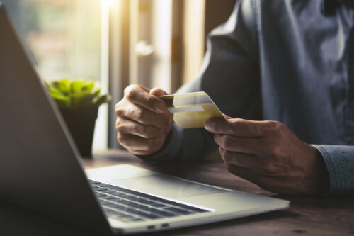 A person in a business casual shirt sits at a wooden desk with a laptop open, holding a credit card, seemingly ready to make an online transfer or conduct a financial transaction.