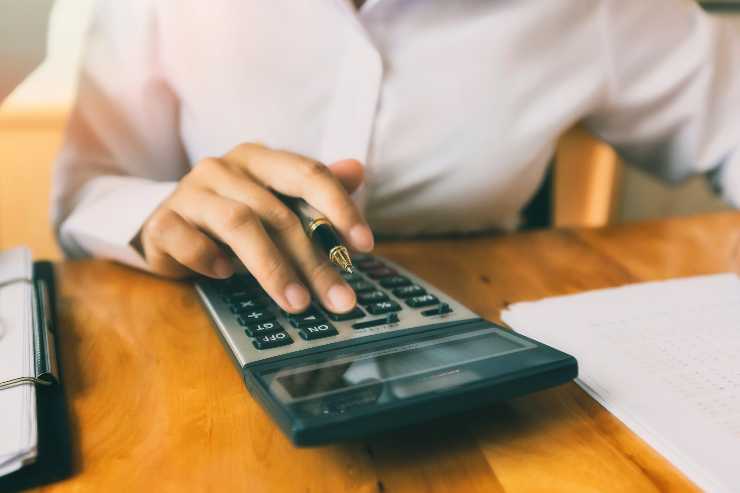 A person in a white shirt using a calculator on a wooden desk with paperwork nearby, possibly managing finances or performing fraudulent accounting tasks.